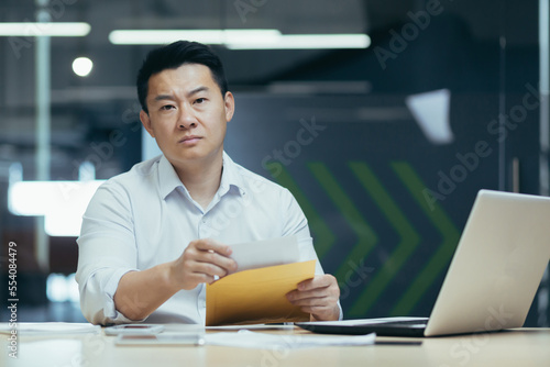 A serious and bespectacled handsome man, an Asian businessman, is sitting in the office at the table, holding an envelope with a letter in his hand. Got bad news, financial problems, bills.