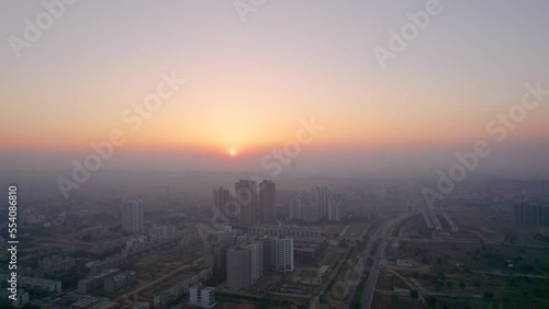 Orbiting aerial drone shot of skyscrapers standing alone in middle of small houses, feilds and busy streets with sunset in distance in the hazy foggy day at gurgaon delhi photo