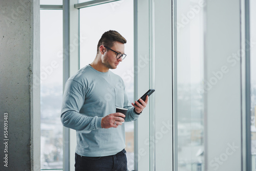 Smiling man in casual clothes standing near window with coffee and looking at smartphone in modern workspace with big window during daytime