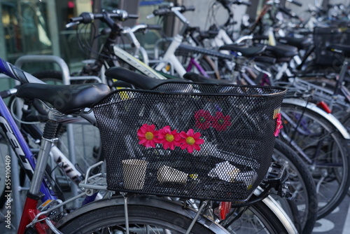 Bicycle with black metal wire basket decorated with red plastic flowers. In the basket there are empty paper coffee cups. A detail of crowded bicycle in parking lot.