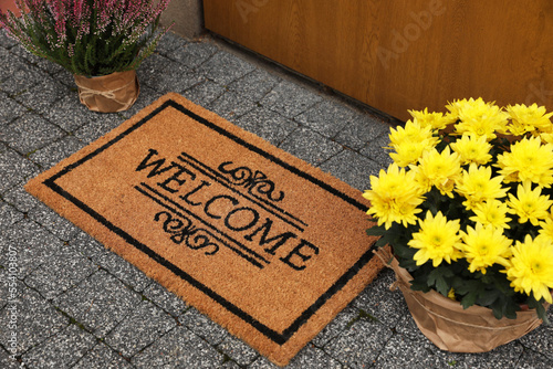 Doormat with word Welcome and beautiful flowers on floor near entrance