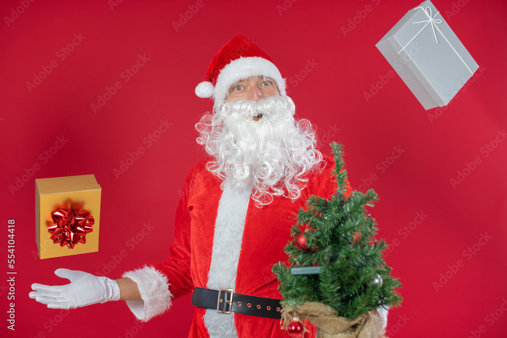 Naklejka premium Portrait von einem verkleideten Weihnachtsmann mit Geschenk und Weihnachtsbaum, isoliert auf roten Hintergrund im Studio