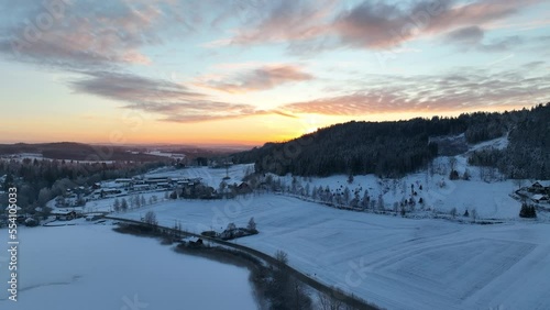 Drone video of a snowy Norwegian landscape in a beautiful winter evening sunset. 4K aerial, slow rise up to see the sun over a hill covered by a forest of pine tree.