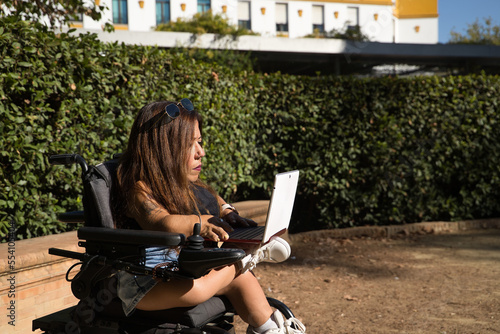 Disabled woman with reduced mobility and small stature in an electric wheelchair working with her laptop computer outdoors. Concept handicap, disability, incapacity, special needs, teleworking. photo