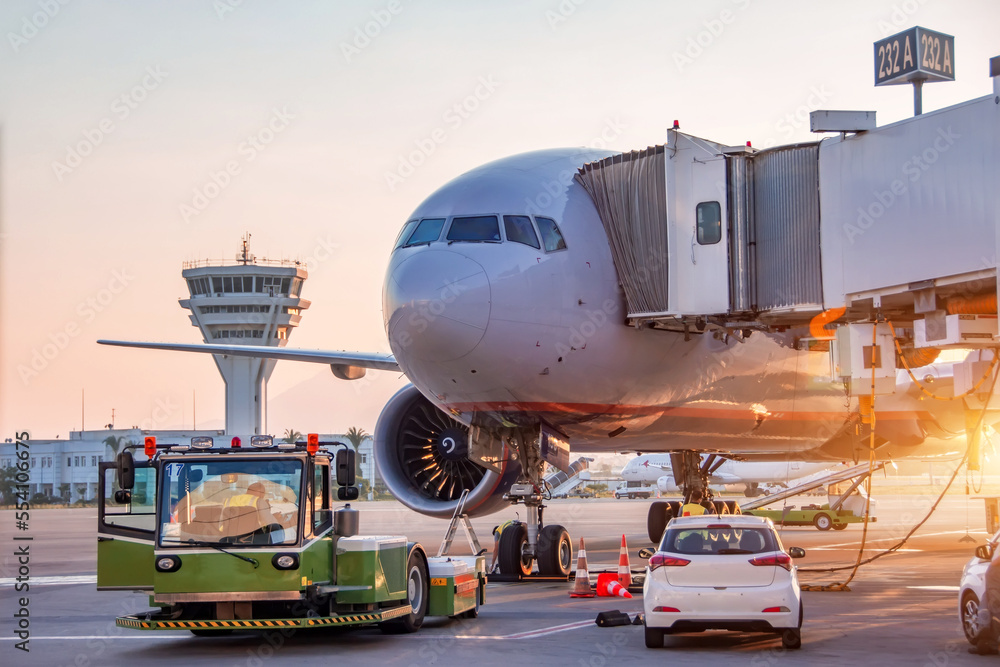 Aircraft is being serviced by airfield ground services at gangway of terminal airport building, preparing for towing and launching into flight in the evening at sunset. Aircraft is being serviced by airfield ground services at gangway of terminal airport building, preparing for towing and launching into flight in the evening at sunset.