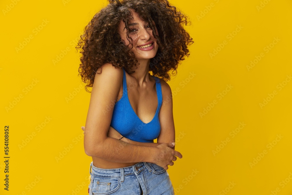 Woman with curly afro hair in a blue t-shirt on. yellow background signs with her hands, look into the camera, smile with teeth and happiness, copy space Woman with curly afro hair in a blue t-shirt on. yellow background signs with her hands, look into the camera, smile with teeth and happiness, copy space