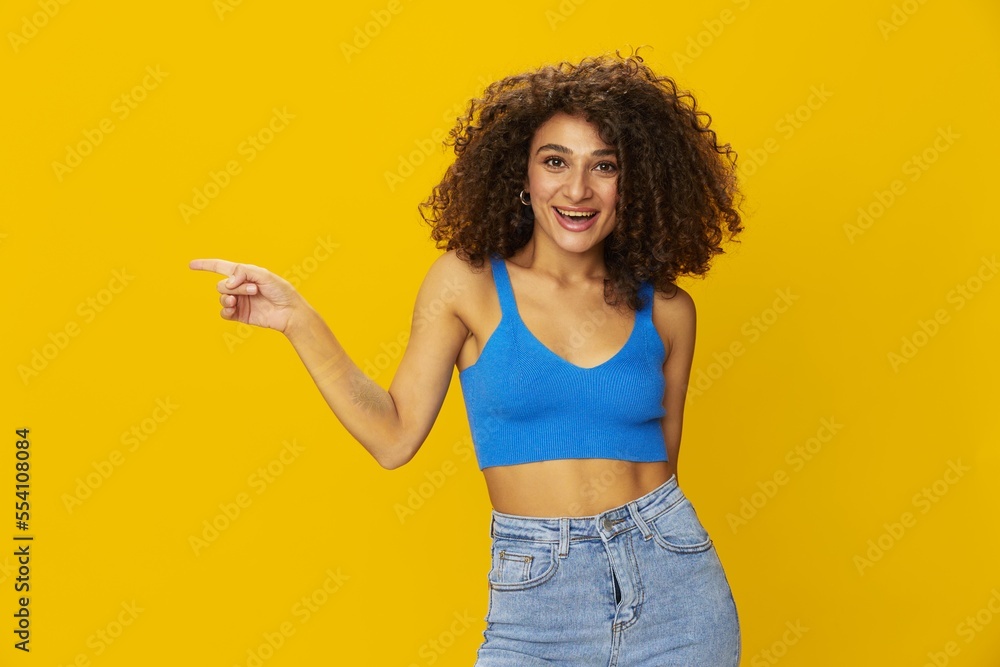 Woman with curly afro hair in a blue t-shirt on. yellow background signs with her hands, look into the camera, smile with teeth and happiness, copy space Woman with curly afro hair in a blue t-shirt on. yellow background signs with her hands, look into the camera, smile with teeth and happiness, copy space