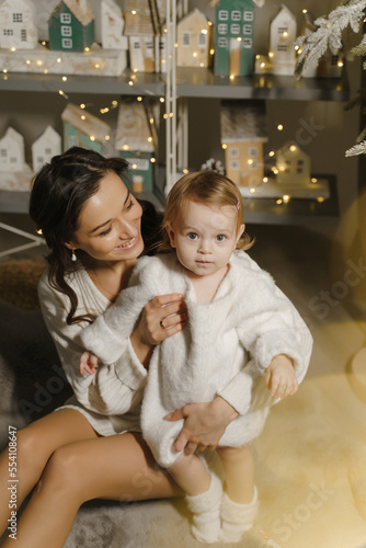 A cute little girl on the background of a Christmas decor. A mother holds a happy child in her arms. Happy holidays, New Year. Cozy warm winter evening at home. Christmas time