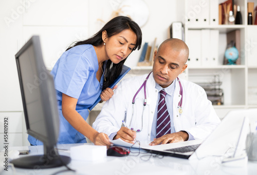 Focused doctor working in medical office  signing papers brought by female assisant