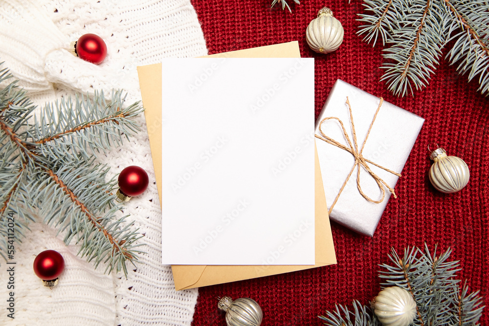Obraz premium Christmas greeting card mockup with envelope, gift box, fir tree branches and holiday decorations on red knitted sweater background, top view, flat lay. White New Year card with winter decor