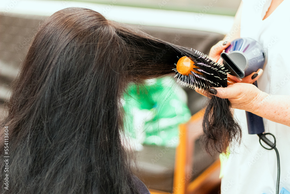 Fototapeta premium A stylist combing a woman's long black hair using a hair dryer and a roller brush