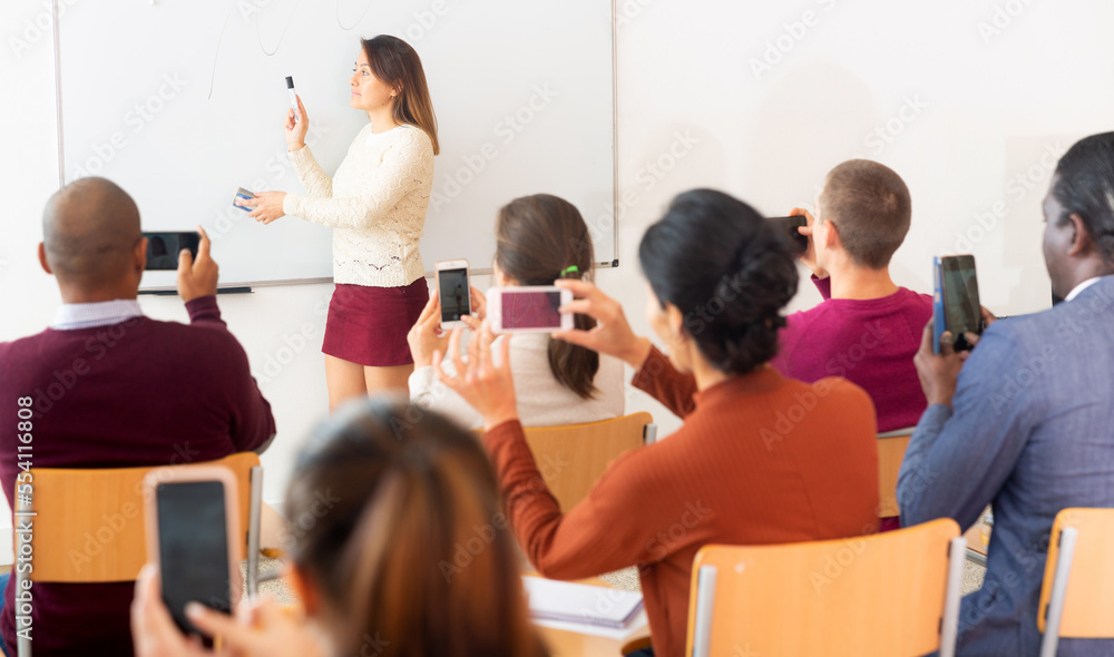 Obraz premium .Female teacher standing in front of whiteboard and people shooting on smartphones her