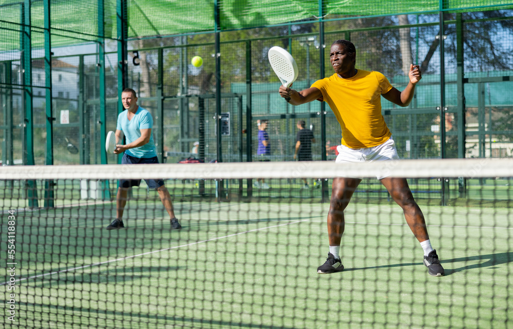 African-american man in sportswear playing padel tennis match with partner during training on court.