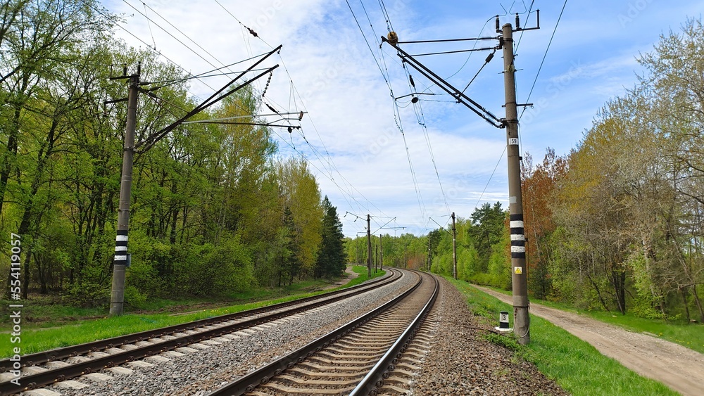 Railway rails are laid on concrete sleepers and sprinkled with crushed stone. The railroad is electrified, along the rails installed concrete poles with wires. Near there are grassy lawns and trees 