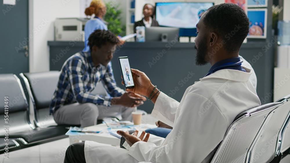 Fototapeta premium Young doctor using video conference on smartphone to talk to specialist about disease treatment, sitting in waiting area. Professional medic chatting on online telemedicine videocall.