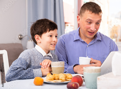 Portrait of young father and son with tea using laptop at table indoors