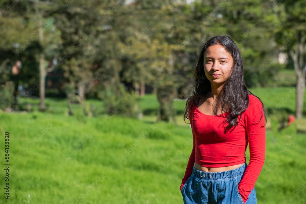 young woman smiling looking at the camera with a red jacket on the side with a nature and forest background young woman smiling looking at the camera with a red jacket on the side with a nature and forest background