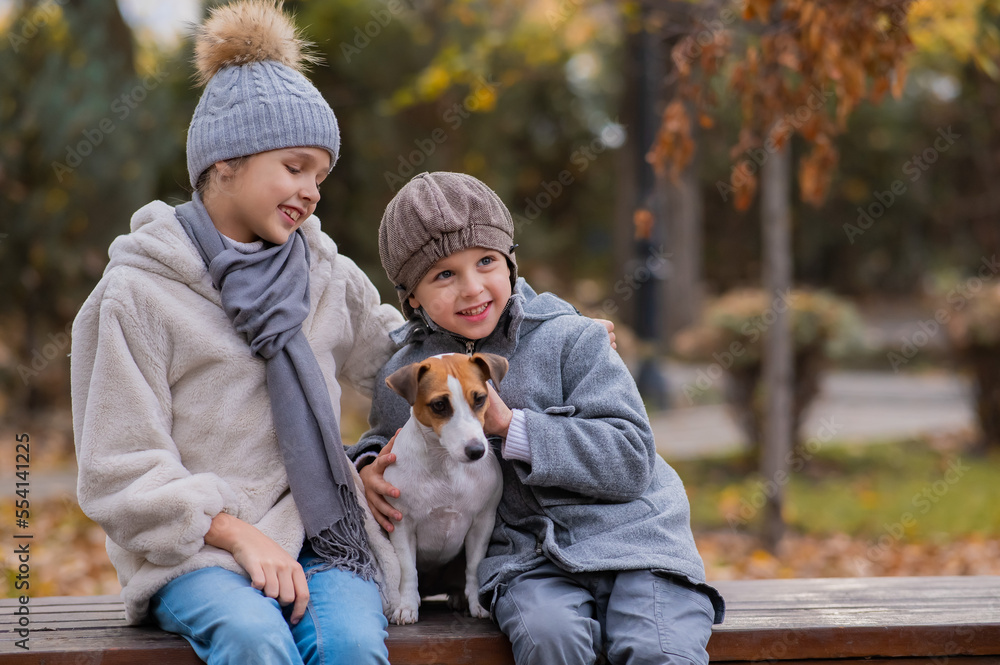 Brother and sister sit in an embrace with a dog on a bench for a walk in the autumn park. Boy, girl and jack russell terrier. 