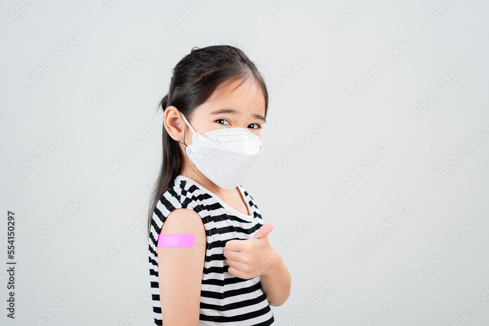 Asian little girl with medical mask showing her arm with bandage after virus vaccine. Kid and child vaccination campaign. Protect your kid with vaccine concept