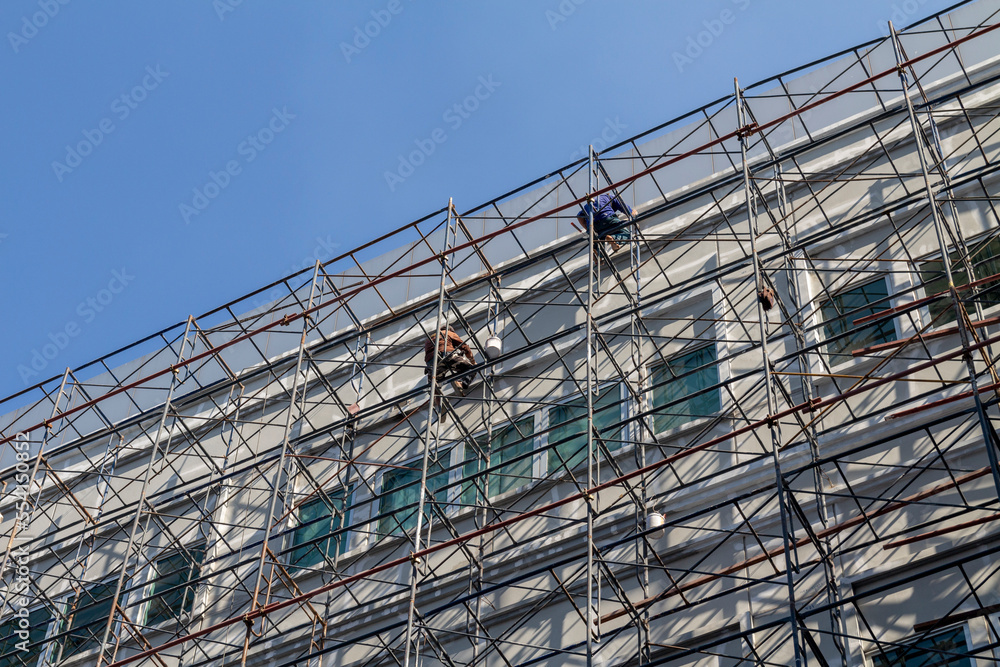 A construction worker is improving the paint of a building by sitting on scaffolding to paint the walls of a building on a high rise.
