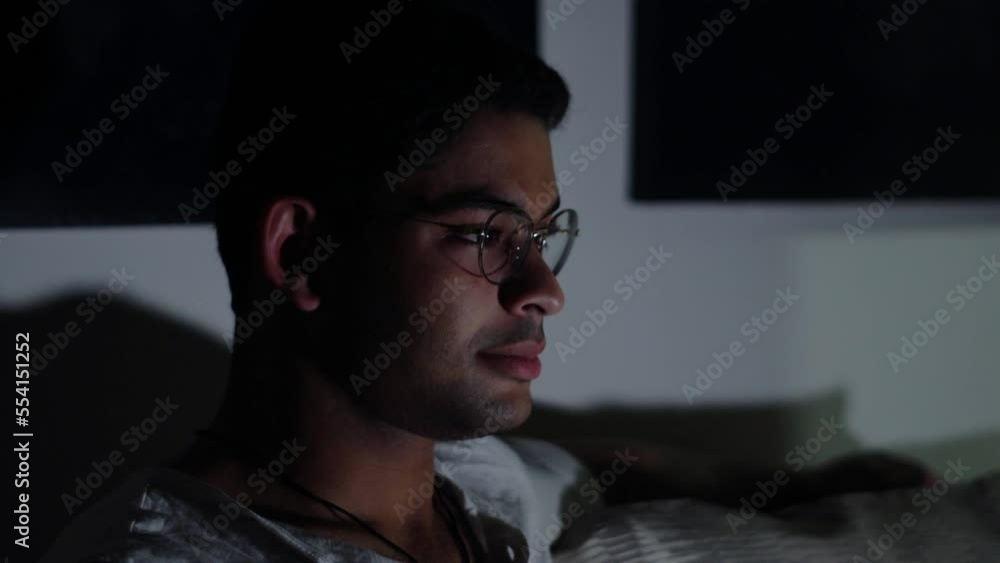 Side View Of A Young Handsome Mixed-Race Indian Man In Eyeglasses Sitting On A Couch, Enjoys Watching Television Alone In A Dark Living Room. - Medium Closeup Side View Of A Young Handsome Mixed-Race Indian Man In Eyeglasses Sitting On A Couch, Enjoys Watching Television Alone In A Dark Living Room. - Medium Closeup