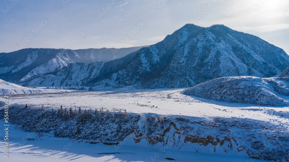 A frozen river in a snowy valley. Coniferous trees grow on the steep banks. A picturesque mountain range against the blue sky. Altai