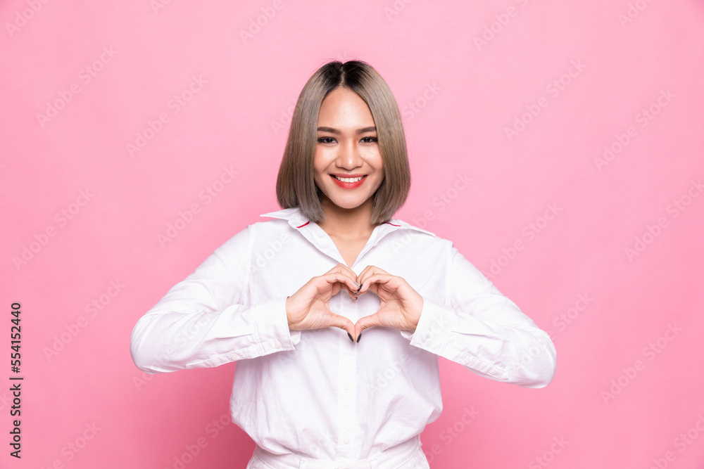 Attractive brunette young Asian woman feels happy and romantic shapes heart gesture expresses tender feelings wears casual white shirt poses against pink background. People affection and care concept Attractive brunette young Asian woman feels happy and romantic shapes heart gesture expresses tender feelings wears casual white shirt poses against pink background. People affection and care concept