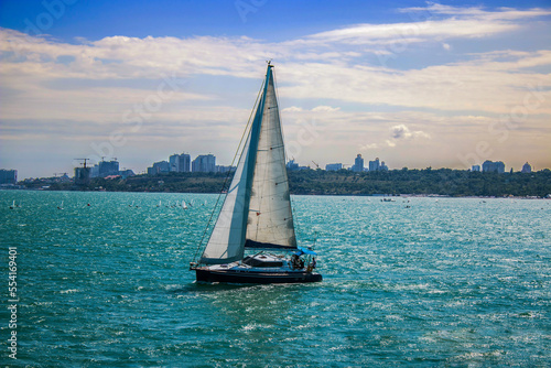 A small beautiful yacht with white sails sails on the blue sea against the background of the resort town