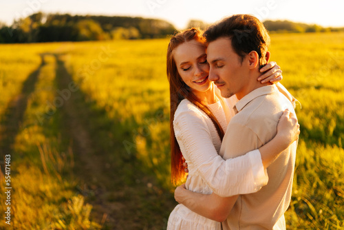 Medium shot portrait of young couple in love with closed eyes standing embracing together on green meadow in summer evening, during sunset with soft sunlight. Concept of romantic date love outdoors.