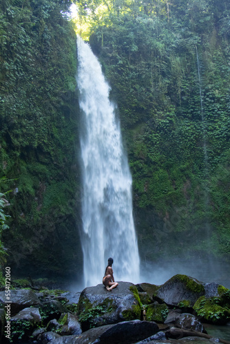Young Asian girl on large rock looking up at massive waterfall in rainforest - Bali, Indonesia