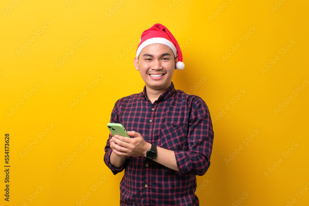 Smiling young Asian man in Santa hat holding smartphone, looking at camera over yellow studio background. Happy New Year 2023 celebration merry holiday concept