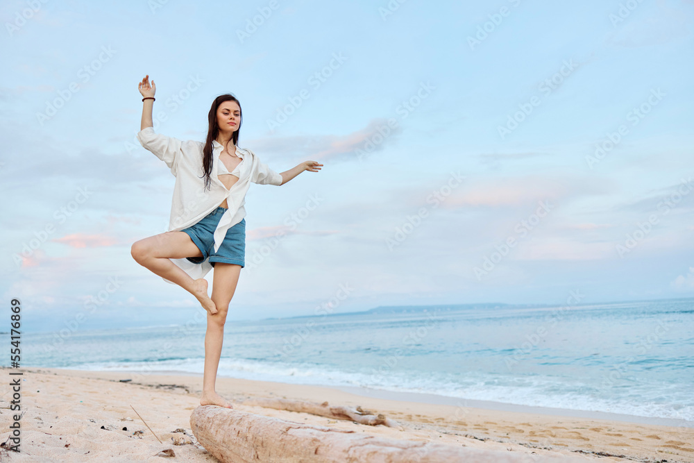 Happy tanned woman in white swimsuit shirt and denim shorts walks on the beach on the sand by the ocean with wet hair after swimming, sunset light and pink clouds in Bali Happy tanned woman in white swimsuit shirt and denim shorts walks on the beach on the sand by the ocean with wet hair after swimming, sunset light and pink clouds in Bali
