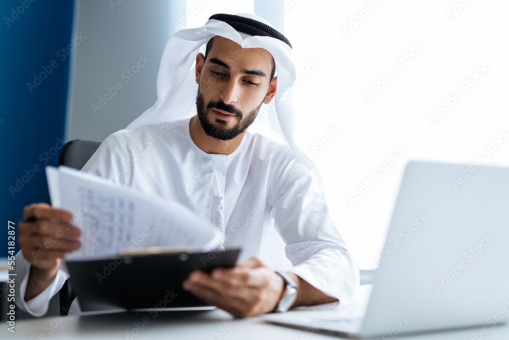 handsome man with dish dasha working in his business office of Dubai. Portraits of a successful businessman in traditional emirates white dress. handsome man with dish dasha working in his business office of Dubai. Portraits of a successful businessman in traditional emirates white dress.