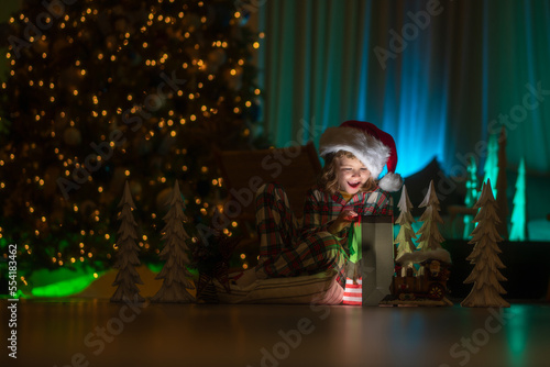 Child opening presents on Xmas eve. Kid in Christmas pajama enjoying winter holiday evening at home near the night Christmas tree.