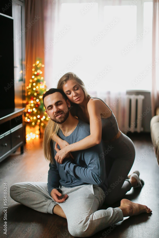 Cozy good Christmas morning. Young couple in grey clothes in living room by Christmas tree. . Man and woman hug at home. Young woman with long hair and big lips in tight tracksuit and young man 