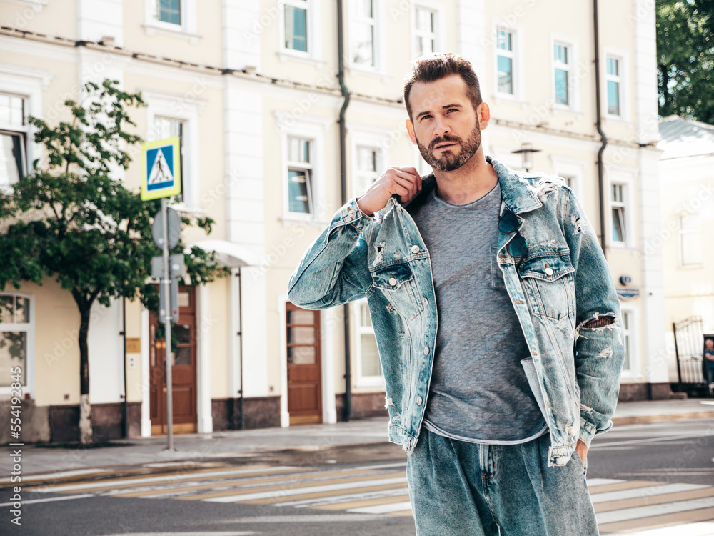 Fototapeta premium Portrait of handsome confident stylish hipster model. Sexy man dressed in jeans jacket. Fashion male posing in the street. In Europe city. Outdoors at sunny day. In sunglasses