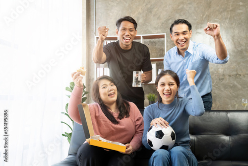 Friends watch sports on TV, cheer and celebrate. Happy diverse asian friend supporters fans sit on couch with popcorn and drinks