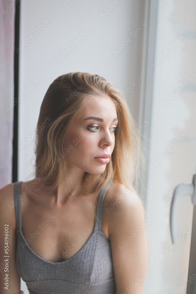 Close-up portrait at window. Caucasian young woman natural blonde with long hair and large silicone lips. Close-up portrait at window. Caucasian young woman natural blonde with long hair and large silicone lips.
