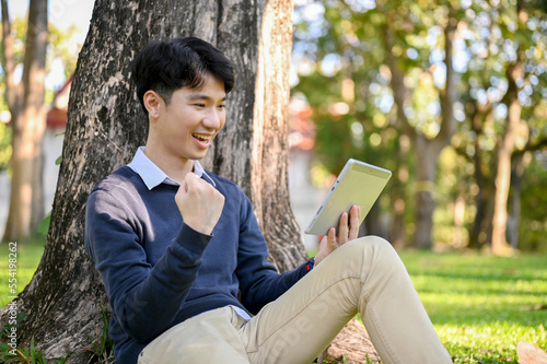 Cheerful Asian male using tablet while sitting under the tree in the park, showing clenched fist