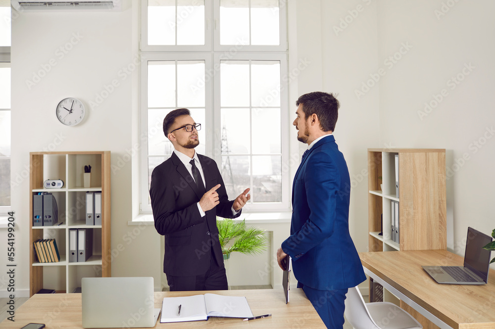 © Studio Romantic - Business people working in the office. Two young men in suits talking while standing by a desk in a modern office interior. Serious manager giving some instructions to a new employee