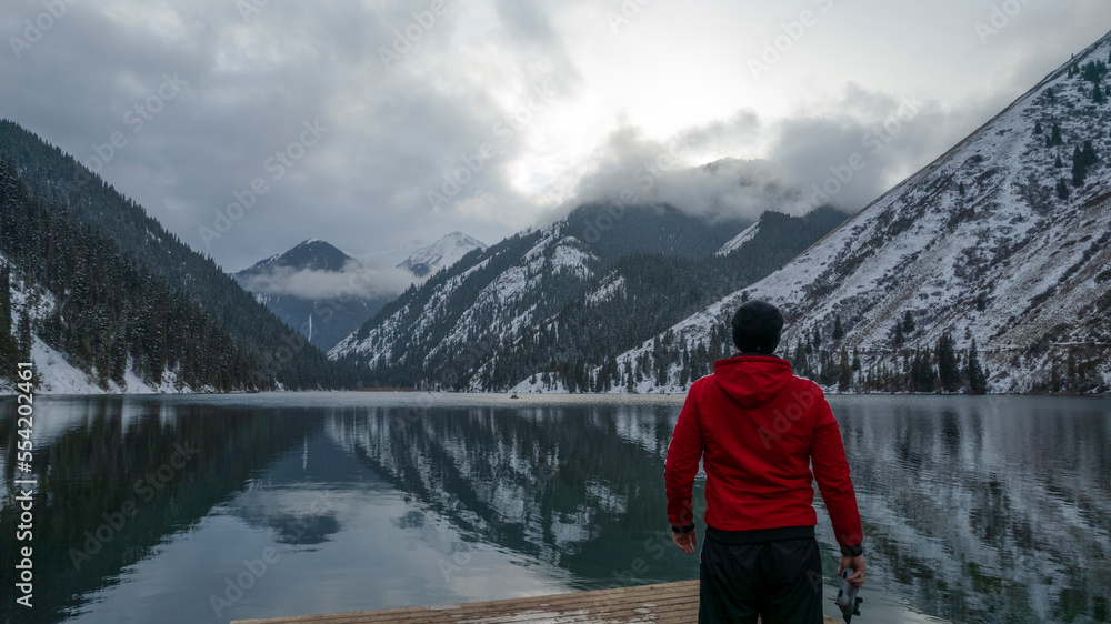 The guy standing on the pier admires the mountain lake. There is a view of the mirrored black color of the water, which reflects snowy mountains, green forest, clouds and a yellow sunset. Kolsai Lake