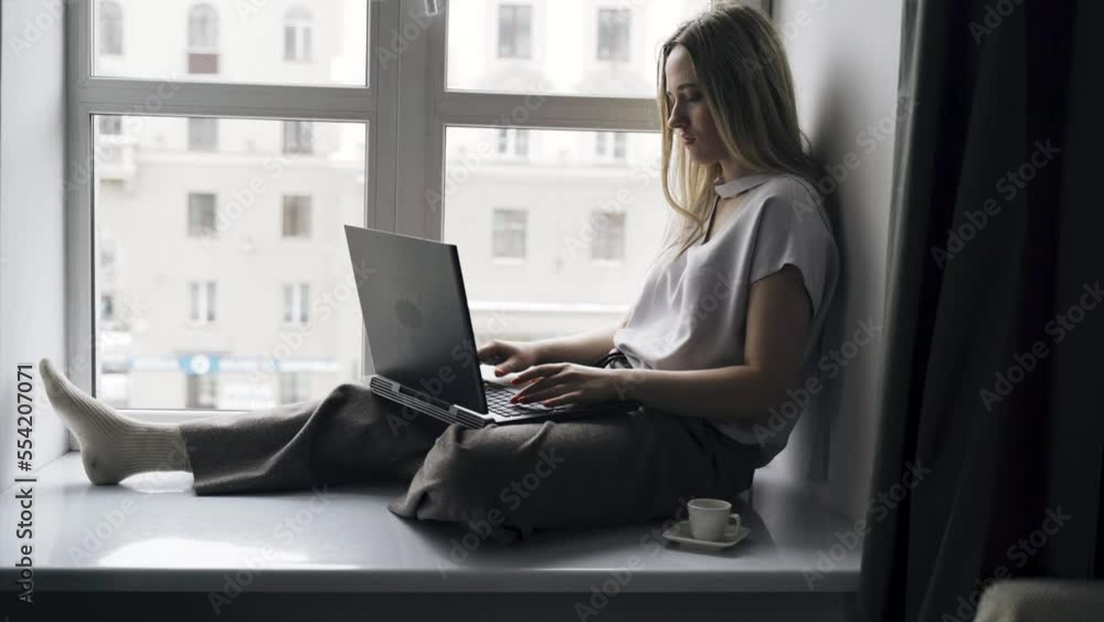 A young woman sits on a windowsill, works on a computer, drinks coffee, looks out the window. Distant work.