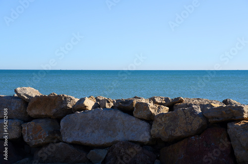Breakwater, Stone wall at sea. Sea with waves against blue sky. Stone pier at sea. Wave breaker at shoreline with stones. Waves in storm breaks on stones at pier. Masonry breakwater in ocean. stone