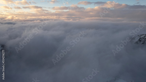 Flying among the ocean of clouds in the mountains. The fog is like big ocean waves crashing against high mountains covered with snow. Spruce trees grow in places. Sunset. Orange rays of the sun