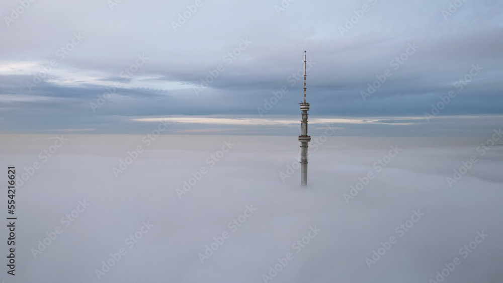 The TV tower looks out of the clouds at sunset. Top view from a drone on a double layer of clouds. Yellow-blue shade of clouds. Shadows on clouds. High mountains are visible in the distance. Kok Tobe