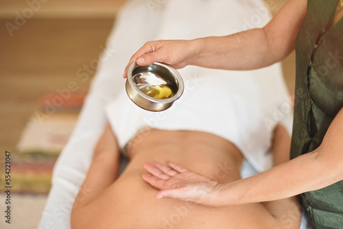 Massage therapist's hands pouring essential oil from bowl on patient's back lying close-up