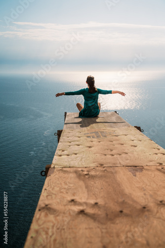 A girl sits on a wooden springboard for jumping with a rope. In a dark green dress with her hands in the air.