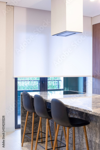 Roller blind large size on the window in the interior. Motorized roller shade white color in the kitchen. Bar counter with built-in hob and hood above it are near the window. Smart home concept. 