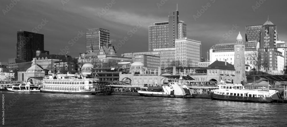 Hamburg panorama with “St Pauli Landungsbrücken“ in the port of the hanseatic metropole in Germany. Black and white wide angle view with waterfront buildings, piers and vessels for harbour cruises.
