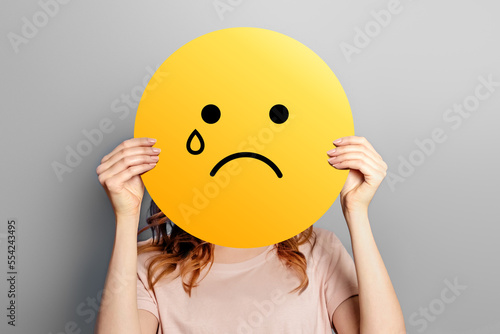 cry smile emoticon symbol. Girl holds a yellow poster with smiley with sad cry face isolated on a grey background